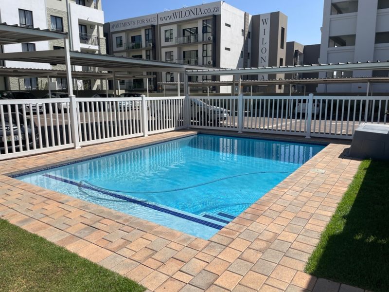 A well-maintained swimming pool area featuring paved surrounds, a white picket fence, and adjacent covered parking structures within an apartment complex.