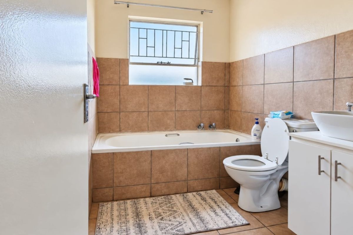 A functional bathroom featuring a built-in bathtub, tiled walls and flooring, a vanity cabinet with a vessel sink, and a window with security bars.