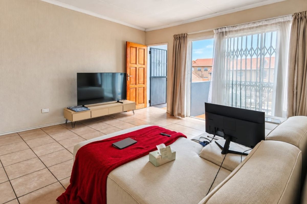 A living room featuring tiled flooring, a sliding glass door with security gate leading to a balcony, and a timber entrance door.