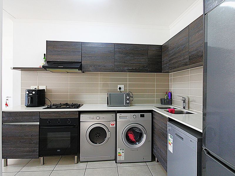 Modern kitchen featuring dark wood-grain cabinetry, stone countertops, tiled backsplash, gas hob, electric oven, and integrated space for laundry appliances.