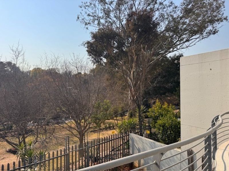 A view from an apartment balcony overlooking a landscaped garden area with mature trees and secure perimeter fencing.