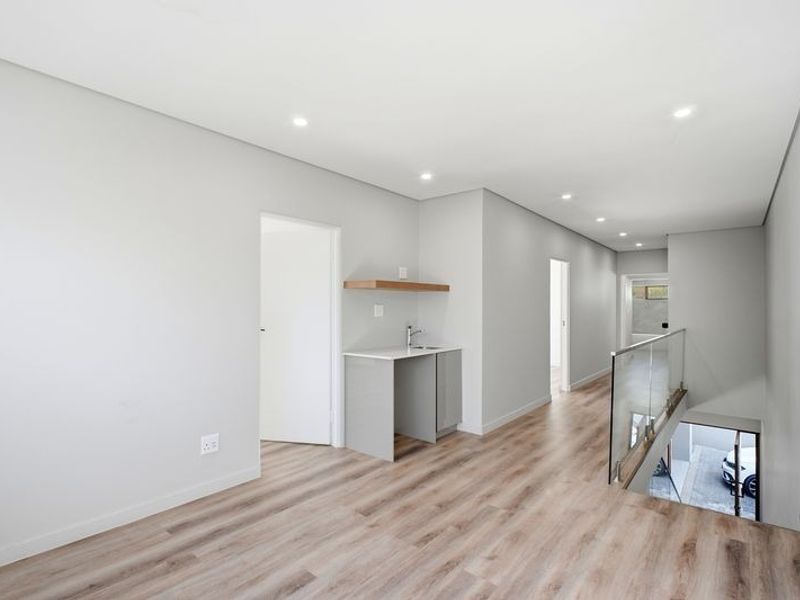 A modern, minimalist hallway featuring light wood-look laminate flooring, recessed lighting, a glass balustrade overlooking the stairs, and a compact built-in kitchenette area.
