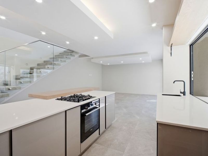 Modern open-plan kitchen featuring stone countertops, integrated gas cooktop, built-in oven, and a sleek glass balustrade staircase in a bright, contemporary space.