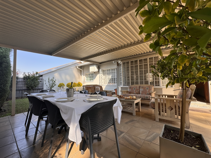A covered outdoor patio featuring tiled flooring, a corrugated metal roof, and a bay window, providing a comfortable space for outdoor dining.