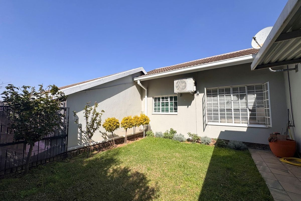 A well-maintained rear garden area featuring a lawn, paved walkway, and exterior wall with security-fitted windows and an air conditioning unit.