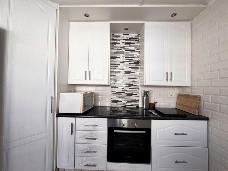 A compact, well-maintained kitchen featuring white cabinetry, granite countertops, a built-in oven, electric hob, and a decorative mosaic tile splashback.