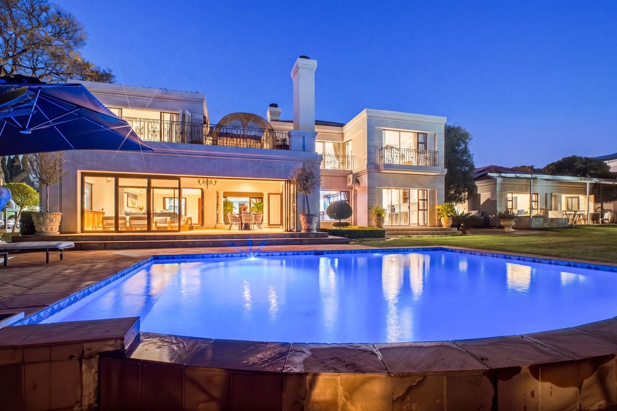 A stunning rear exterior view of a luxury residence featuring a large illuminated swimming pool, expansive patio, and multi-level architectural design at dusk.