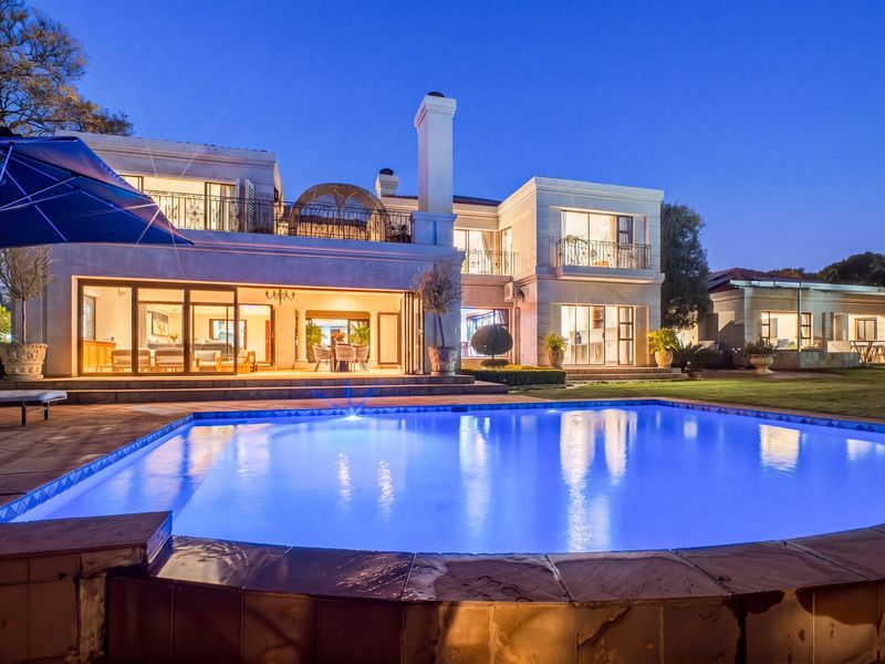 A stunning rear exterior view of a luxury residence featuring a large illuminated swimming pool, expansive patio, and multi-level architectural design at dusk.