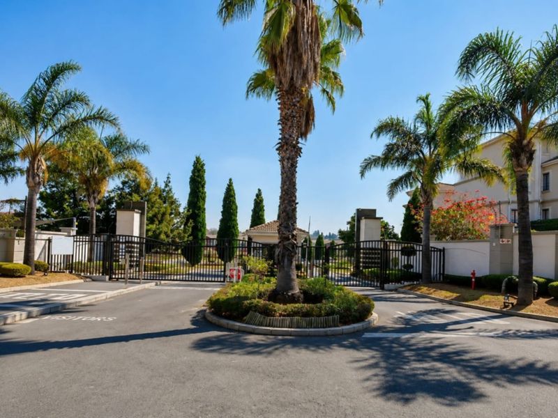 The main entrance to the apartment complex featuring a secure gated access point, paved driveway, and landscaped traffic island with mature palm trees.
