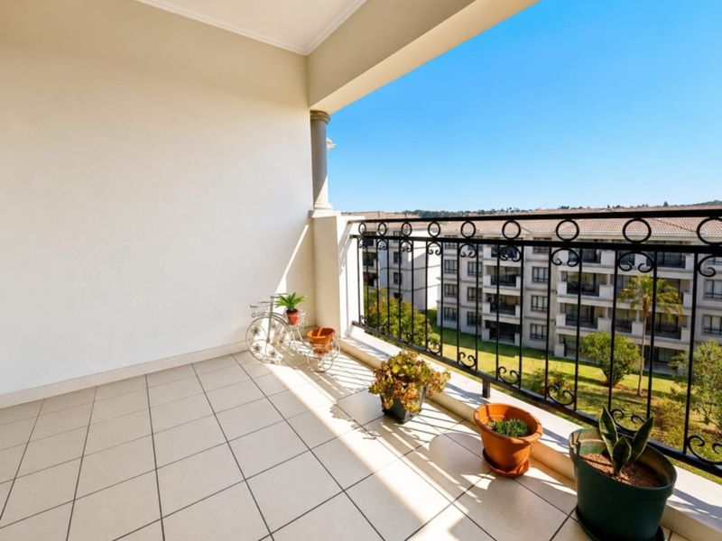 A spacious, covered balcony featuring tiled flooring and a decorative wrought iron balustrade, offering an open view of the surrounding apartment complex.