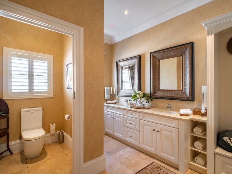 Spacious bathroom featuring a double vanity with dual sinks, built-in cabinetry, stone flooring, and a separate toilet area with plantation shutters.