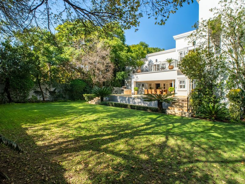A spacious rear garden featuring a well-maintained lawn, mature trees, and a covered patio area with an upper-level balcony overlooking the yard.
