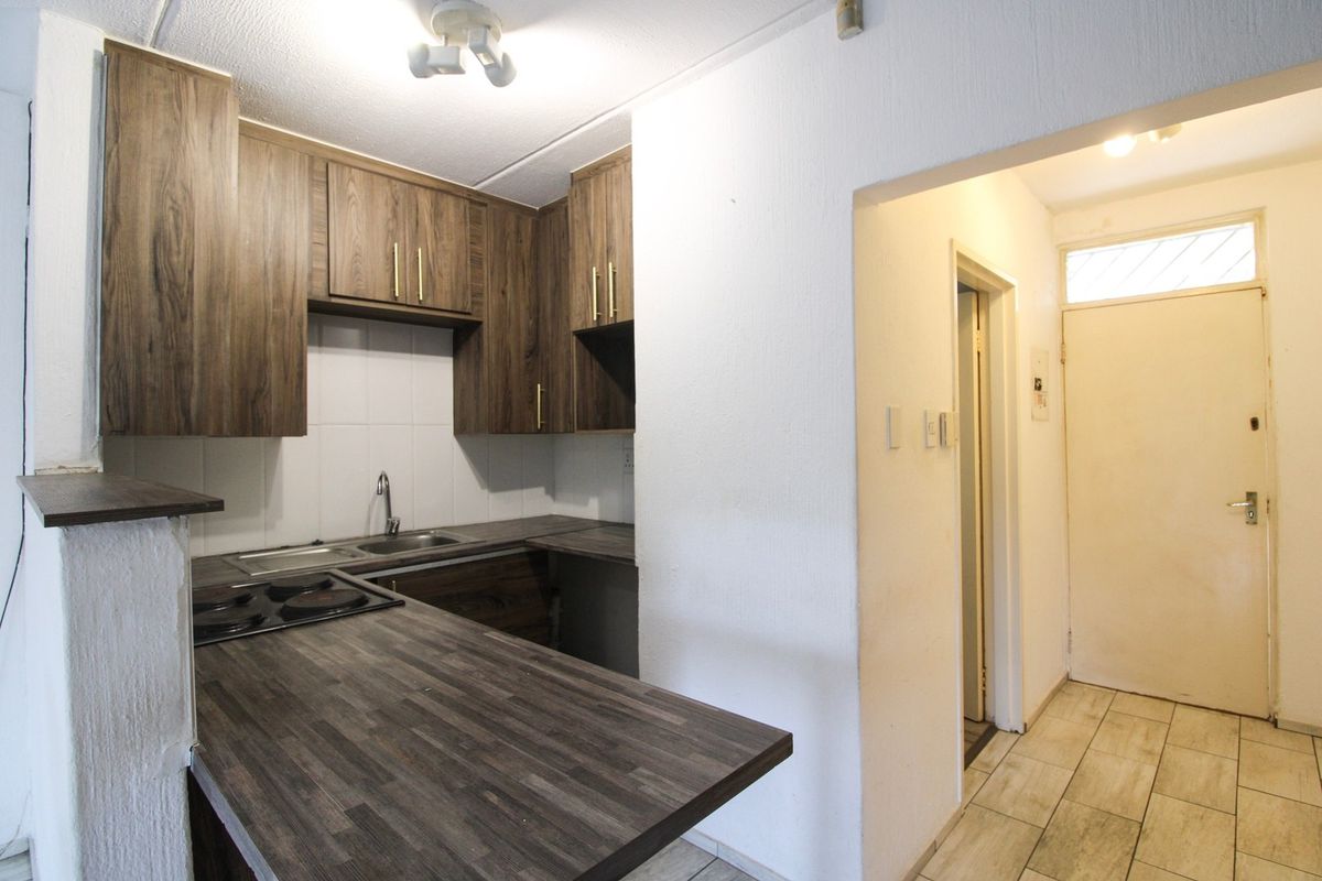 A compact kitchen featuring wood-look laminate countertops, built-in cabinetry, and an integrated electric hob, adjacent to the apartment entrance hallway.