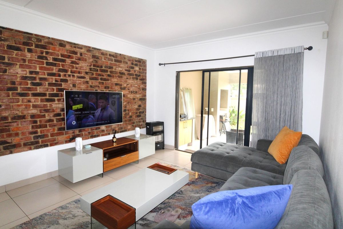 A well-lit living room featuring an exposed brick accent wall, tiled flooring, and large sliding glass doors providing access to the patio area.