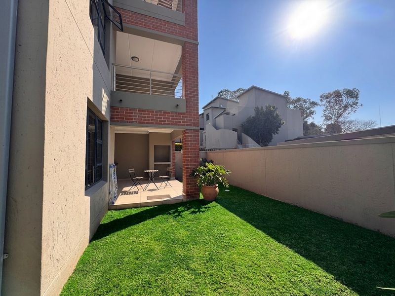 Rear view of the apartment building featuring a covered patio, balcony, and a private garden area with artificial lawn and boundary wall.