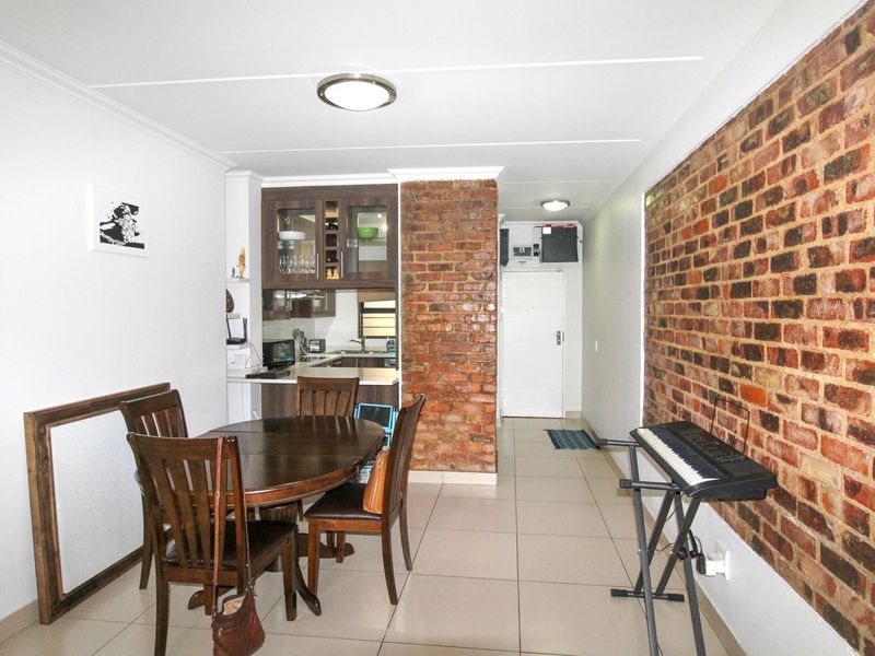 Open plan living and dining area featuring tiled floors and an exposed brick feature wall, leading into a compact kitchen space.