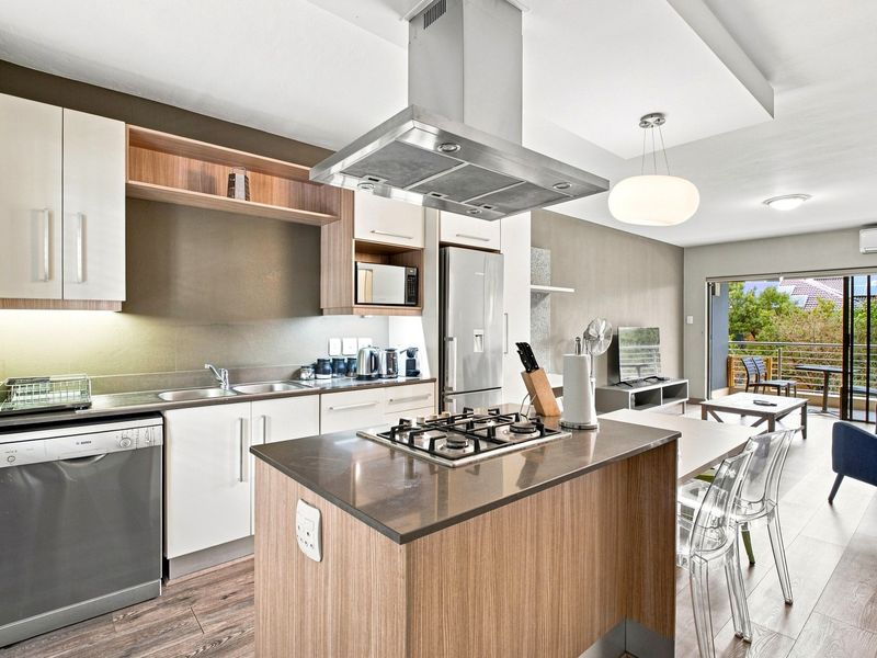 Modern open-plan kitchen featuring a central island with gas hob, stainless steel extractor, and integrated appliances, flowing into a bright living area with balcony access.