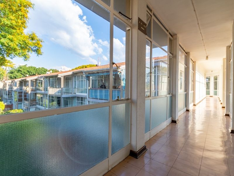 A bright, open-air style apartment hallway featuring tiled flooring and large glass windows overlooking the building exterior.