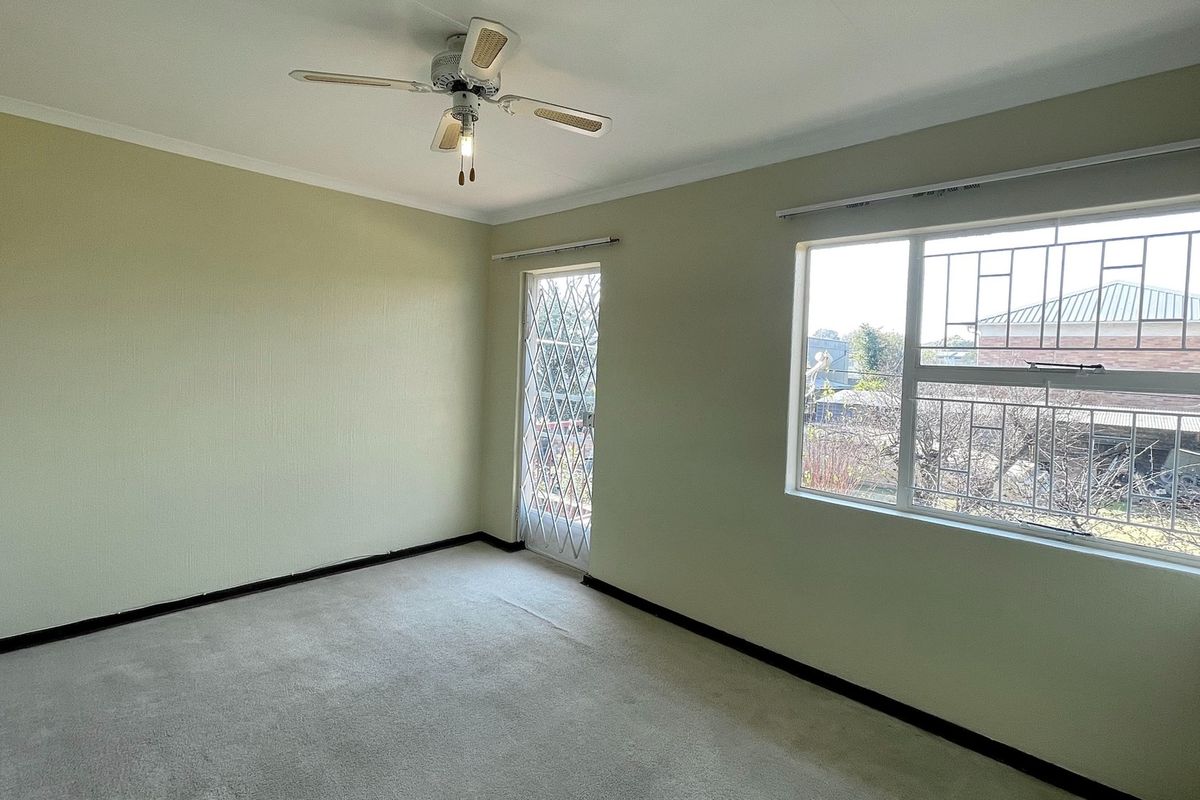 A spacious, empty bedroom featuring neutral walls, carpeted flooring, a ceiling fan, and a security gate leading to the exterior.