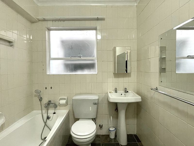 A dated bathroom featuring a standard bathtub, pedestal basin, and toilet, finished with full-height wall tiling and a frosted window for privacy.