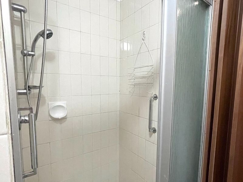 A functional shower enclosure featuring white wall tiles, a handheld shower head, and a safety grab rail.