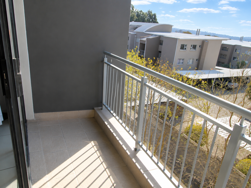 A clean, modern balcony featuring tiled flooring and a secure metal balustrade, offering an elevated view of the surrounding apartment complex.