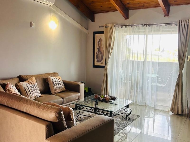 A well-maintained living room featuring high exposed timber beam ceilings, polished tile flooring, and direct access to a balcony through sliding glass doors.