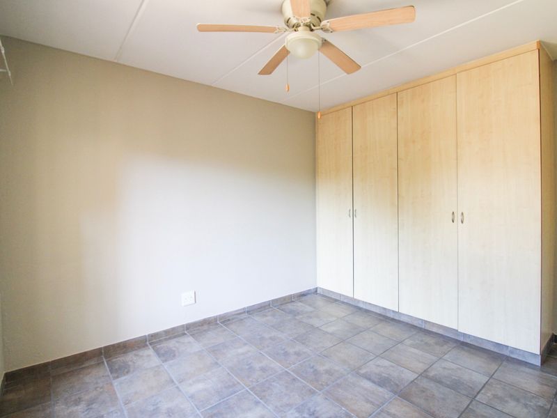 A clean, empty bedroom featuring neutral tiled flooring, built-in wooden cupboards, and a ceiling fan.