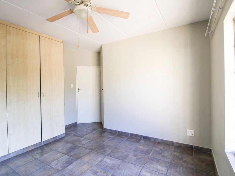 A bright, empty bedroom featuring neutral tiled flooring, built-in wooden cupboards, and a ceiling fan, offering a clean canvas for personalization.