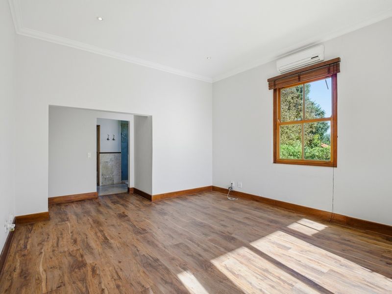 A bright, empty bedroom featuring laminate flooring, timber-framed windows, and a wall-mounted air conditioning unit, with an adjoining space visible.