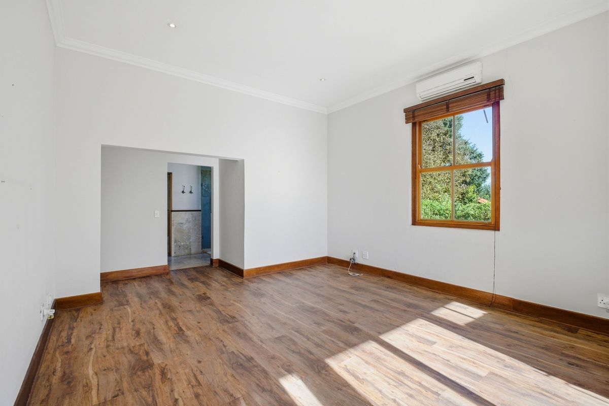 A bright, empty bedroom featuring laminate flooring, timber-framed windows, and a wall-mounted air conditioning unit, with an adjoining space visible.