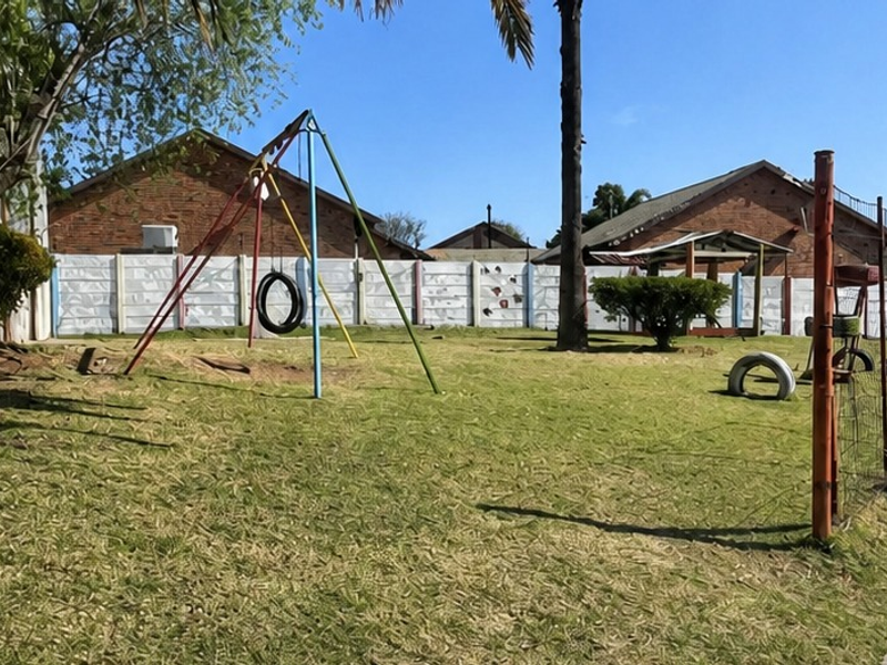 A communal garden area featuring a lawn, mature trees, a precast concrete boundary wall, and metal perimeter fencing with an adjacent paved walkway.