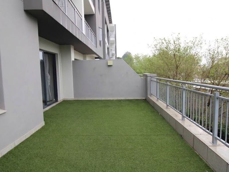 A spacious apartment balcony featuring artificial grass flooring, a secure metal balustrade, and direct access through sliding glass doors.