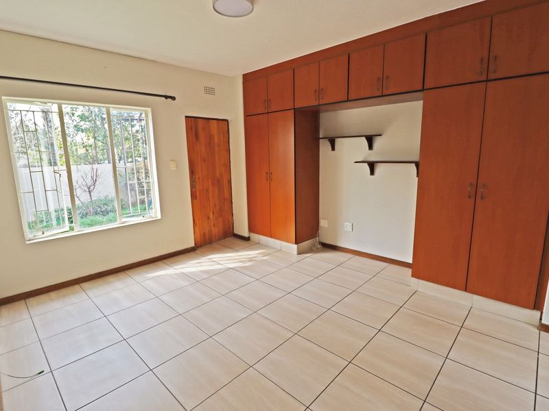 A bright, empty bedroom featuring tiled flooring, extensive built-in wooden cabinetry, and a window with security bars overlooking the garden.