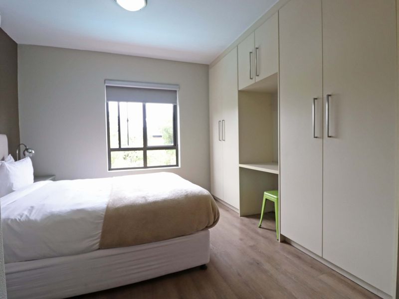 A well-maintained bedroom featuring neutral laminate flooring, a large window with a roller blind, and extensive built-in cabinetry with an integrated desk space.