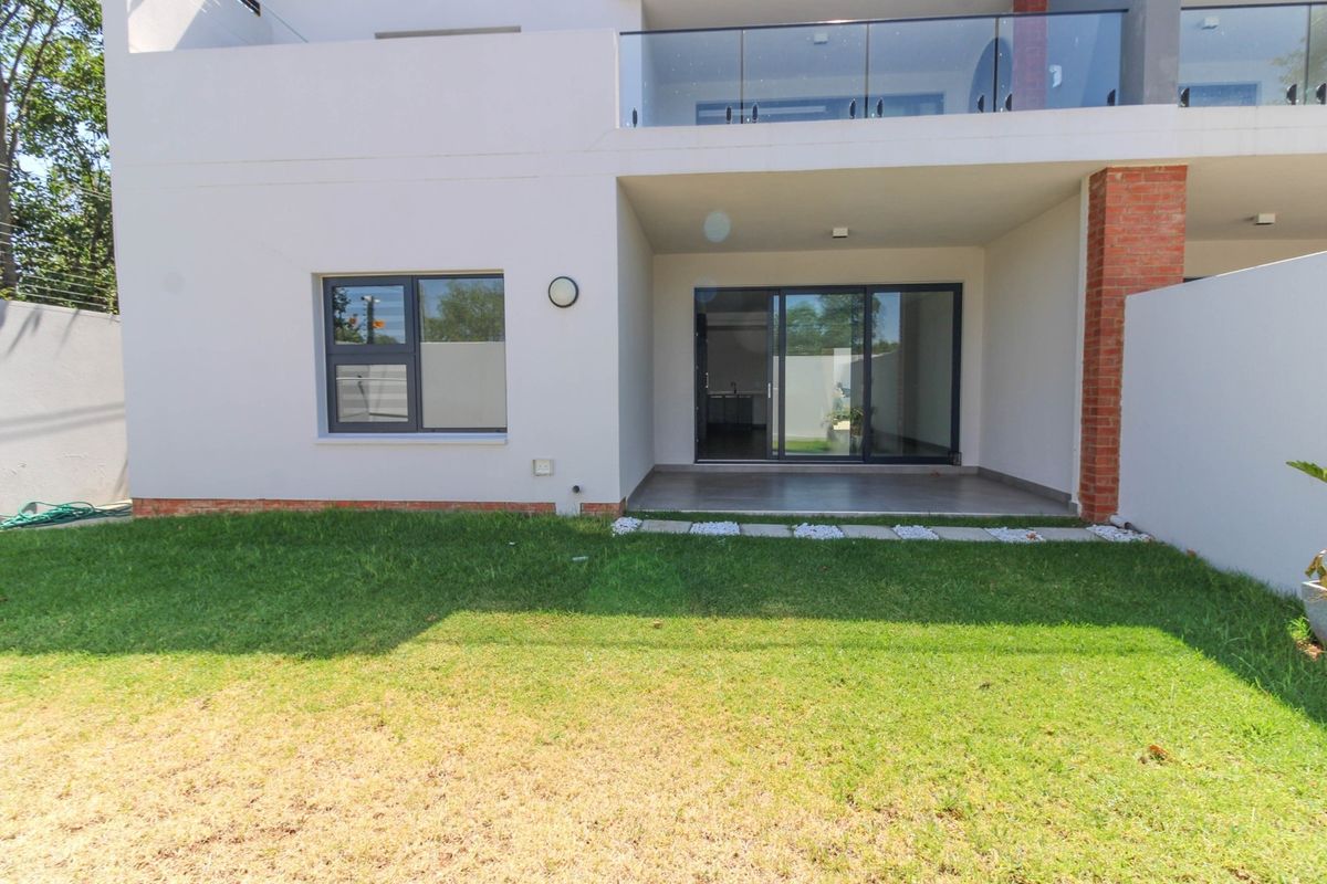 Rear view of the apartment featuring a covered patio, glass sliding doors, and a private garden area with a glass-railed balcony above.