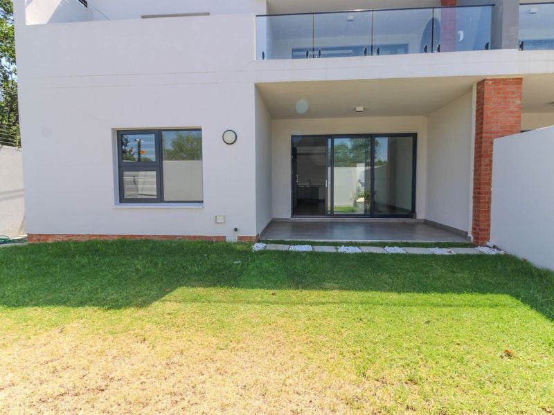 Rear view of the apartment featuring a covered patio, glass sliding doors, and a private garden area with a glass-railed balcony above.
