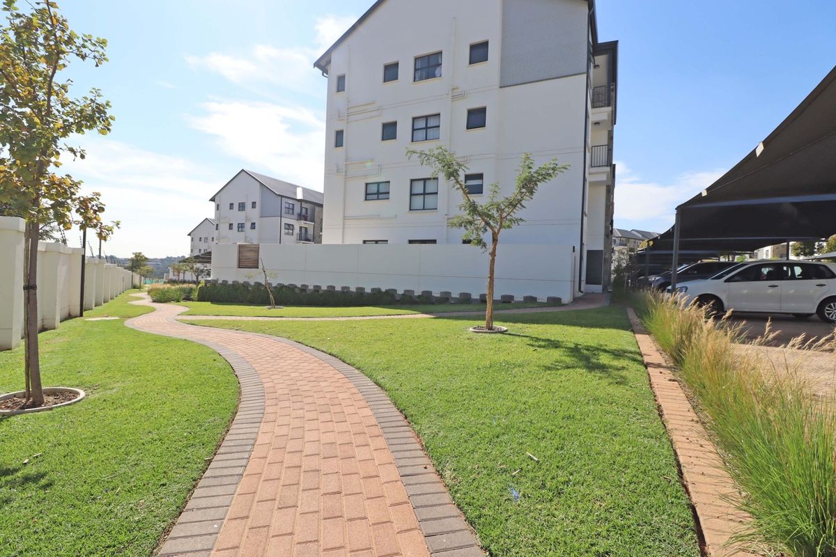 A well-maintained apartment complex exterior featuring a paved walkway, landscaped lawn, and covered parking bays for residents.