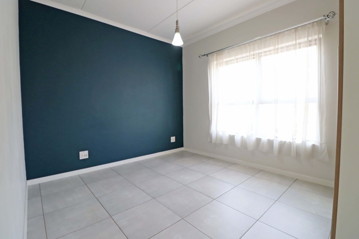 A bright, empty bedroom featuring neutral tiled flooring, a striking dark blue accent wall, and a large window with a fitted curtain rod.