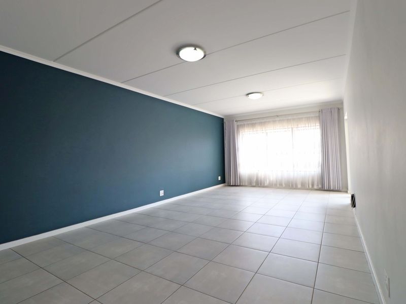 A spacious, empty living room featuring neutral tiled flooring, a bold blue accent wall, and ample natural light from the large window.
