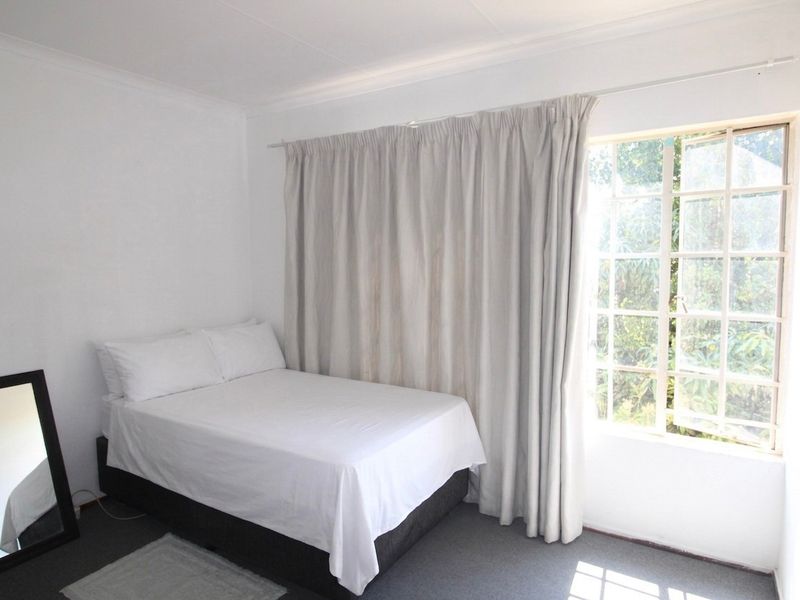 A simple bedroom featuring neutral walls, carpeted flooring, and a large window with curtains allowing natural light.