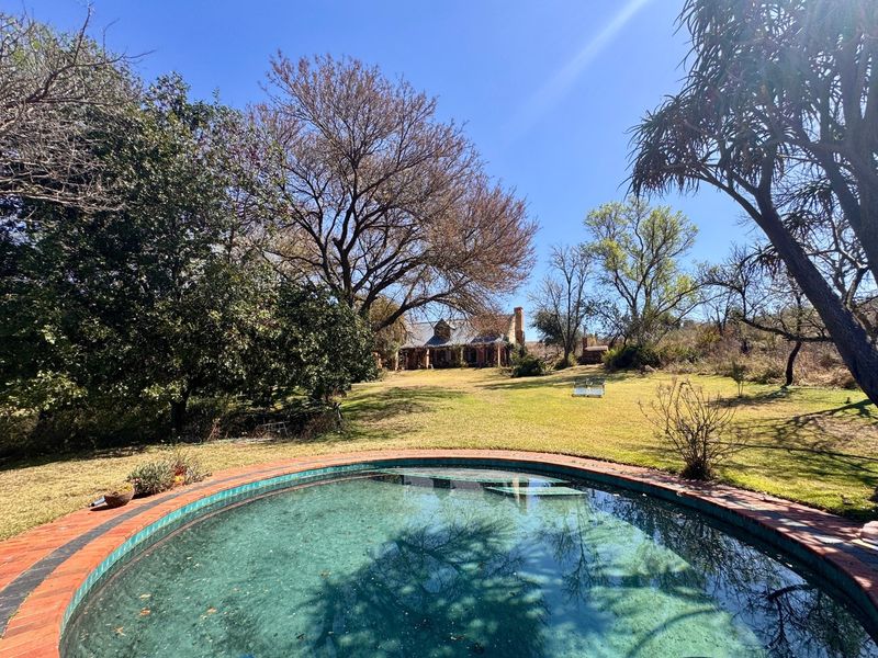 A view of a circular swimming pool with brick coping, set within a spacious garden with mature trees and the main dwelling in the background.