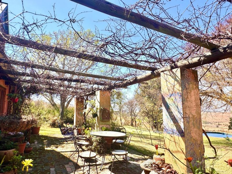 A rustic outdoor patio area featuring a wooden pergola structure, brick paving, and masonry pillars overlooking the garden and surrounding landscape.