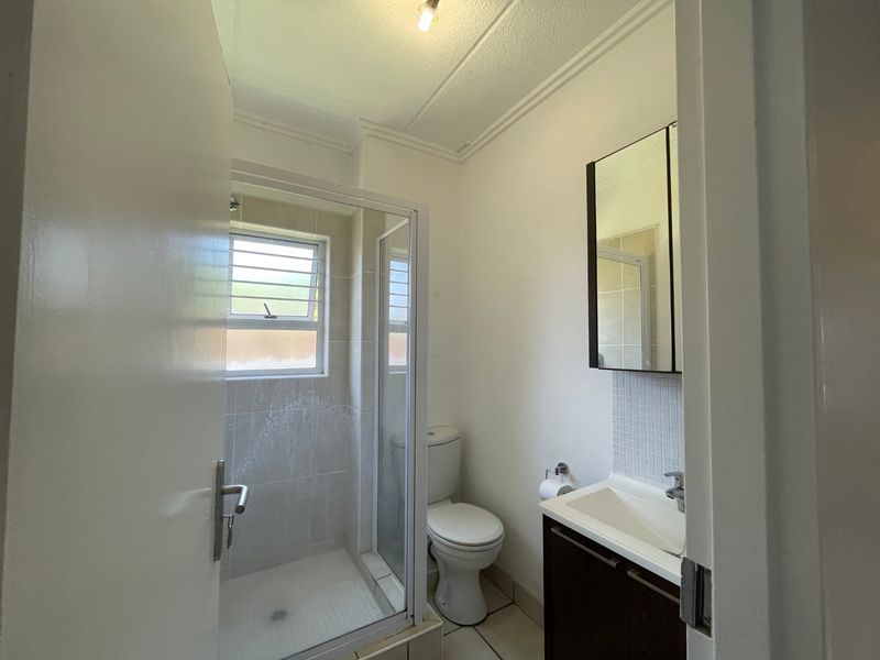 A functional bathroom featuring a shower enclosure, toilet, and vanity cabinet with a mirror, finished with tiled flooring and a louvered window.