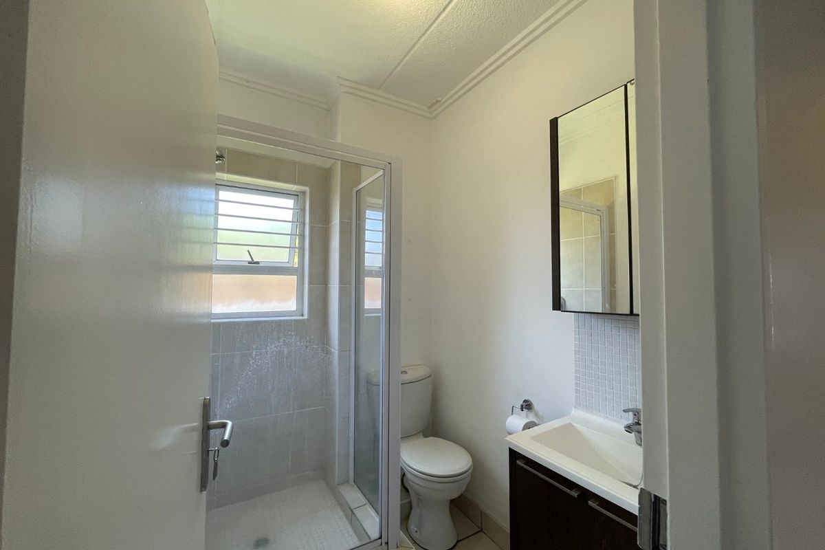 A functional bathroom featuring a shower enclosure, toilet, and vanity cabinet with a mirror, finished with tiled flooring and a louvered window.