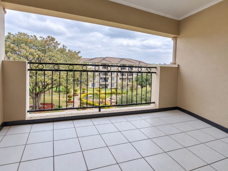 A spacious covered balcony featuring tiled flooring and a decorative wrought iron balustrade, offering pleasant views of the surrounding estate gardens.