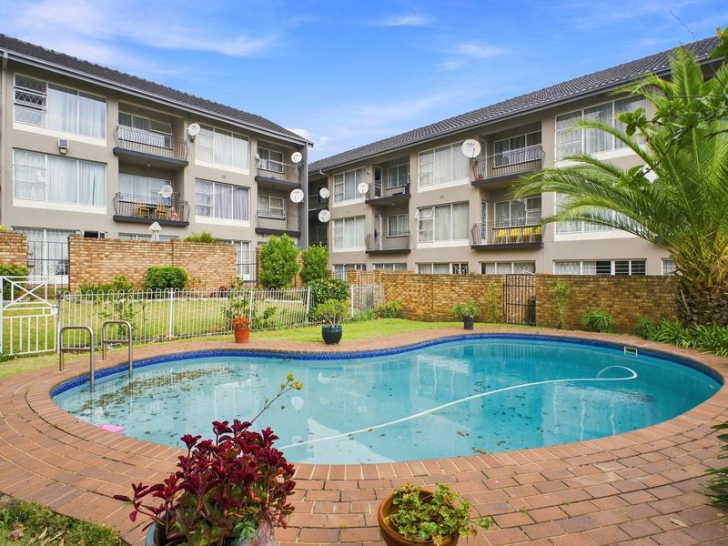 Communal apartment complex pool area featuring a kidney-shaped swimming pool, brick paving, and landscaped garden borders with surrounding residential balconies.