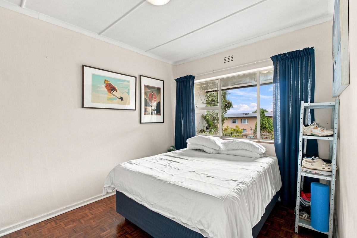 A bedroom featuring parquet flooring, a steel-framed window with security bars, and a ceiling vent, offering a functional space with natural light.