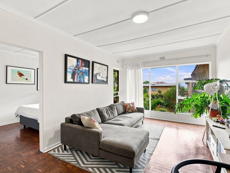 Bright living room featuring classic parquet flooring, large windows providing natural light, and a clean, neutral aesthetic with an open flow to the bedroom.