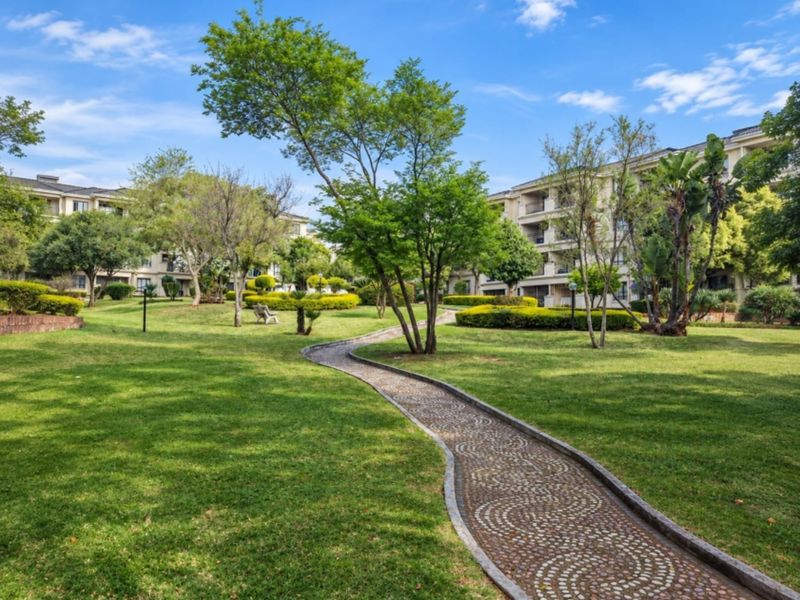 A well-maintained communal garden area featuring a winding pebble pathway, lush green lawns, and mature trees within an apartment complex.